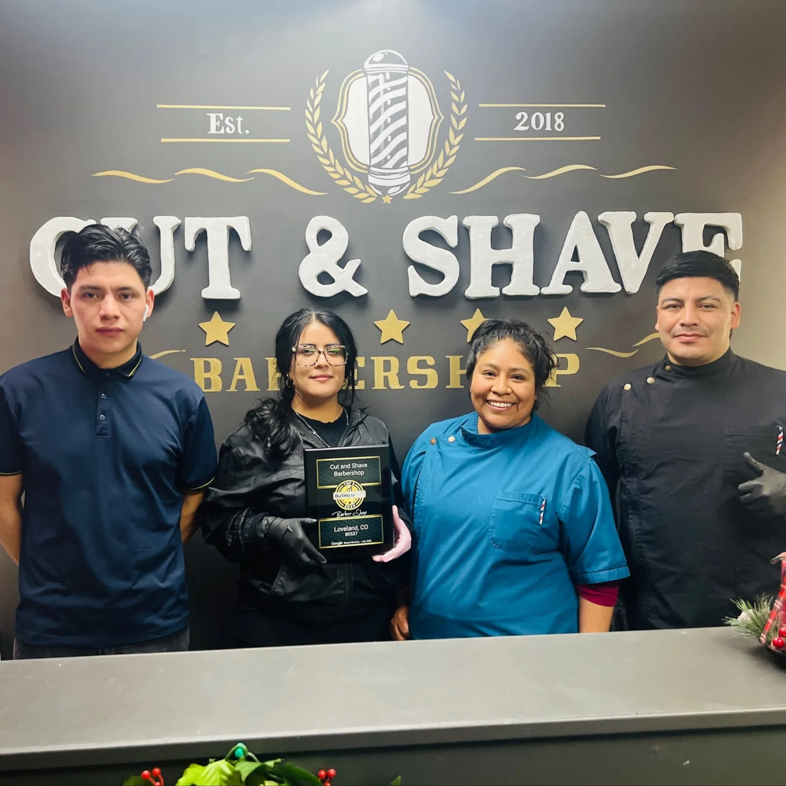 Four members of the Cut and Shave team standing in front of the shop's lit Cut & Shave Barbershop sign, one holding a plaque